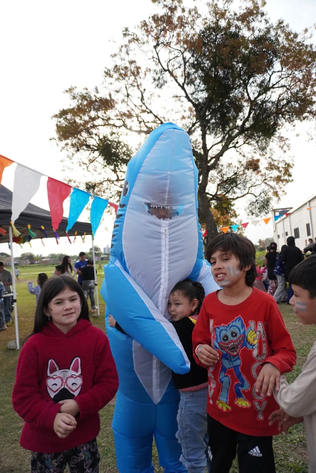 Chicos y chicas junto a una persona con un disfraz de tiburón azul, al aire libre.